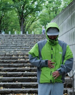 ゴールドウインオンラインストア公式 | STAFF PICKS AUTUMN OUTERWEAR STYLE | ライムグリーン×グレーの防水シェルジャケットを着用し、フードを被った状態でフロントジップ周辺の仕様を確認できるアウトドアルック。